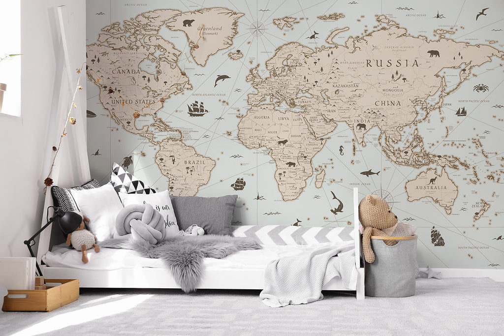 A serene room featuring the Explorer Atlas, World Map Wallpaper in Blue on the main wall. The detailed map displays continents and countries in neutral tones. A cozy bed with striped bedding is in the foreground, accompanied by a teddy bear and toys.