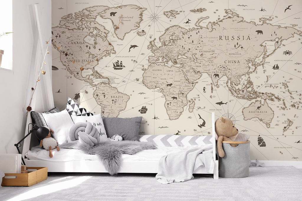 A serene room featuring the Explorer Atlas, World Map Wallpaper in Sand on the main wall. The detailed map displays continents and countries in neutral tones. A cozy bed with striped bedding is in the foreground, accompanied by a teddy bear and toys.
