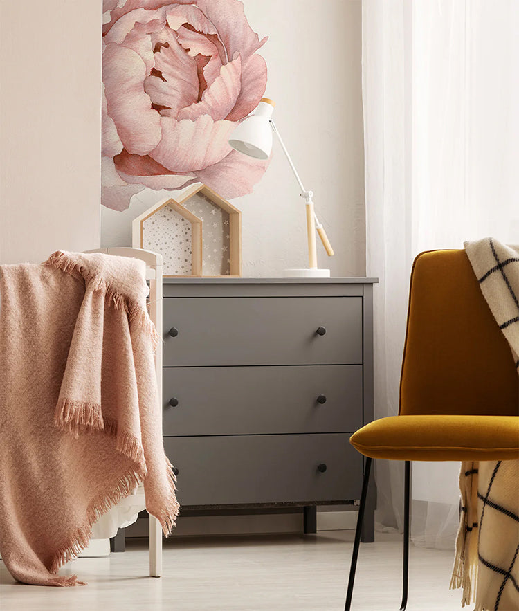 A cozy room featuring the Extra Large Solo Peony, Wall Decals in Blush. A gray dresser with a white lamp, wooden decor, and a draped pink throw, next to a yellow chair with a checkered throw.