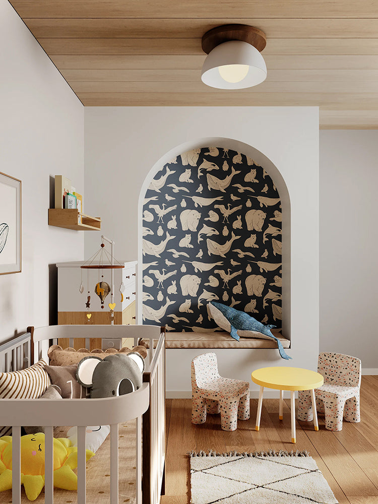 A modern nursery with wooden flooring, a white crib, matching dresser, and shelves filled with toys and books. A whimsical whale illustration adorns the wall. The room is accented by a round yellow table, and two small chairs with animal motifs. The niche features the Frosty Friends, Animal Pattern Wallpaper in Blue.