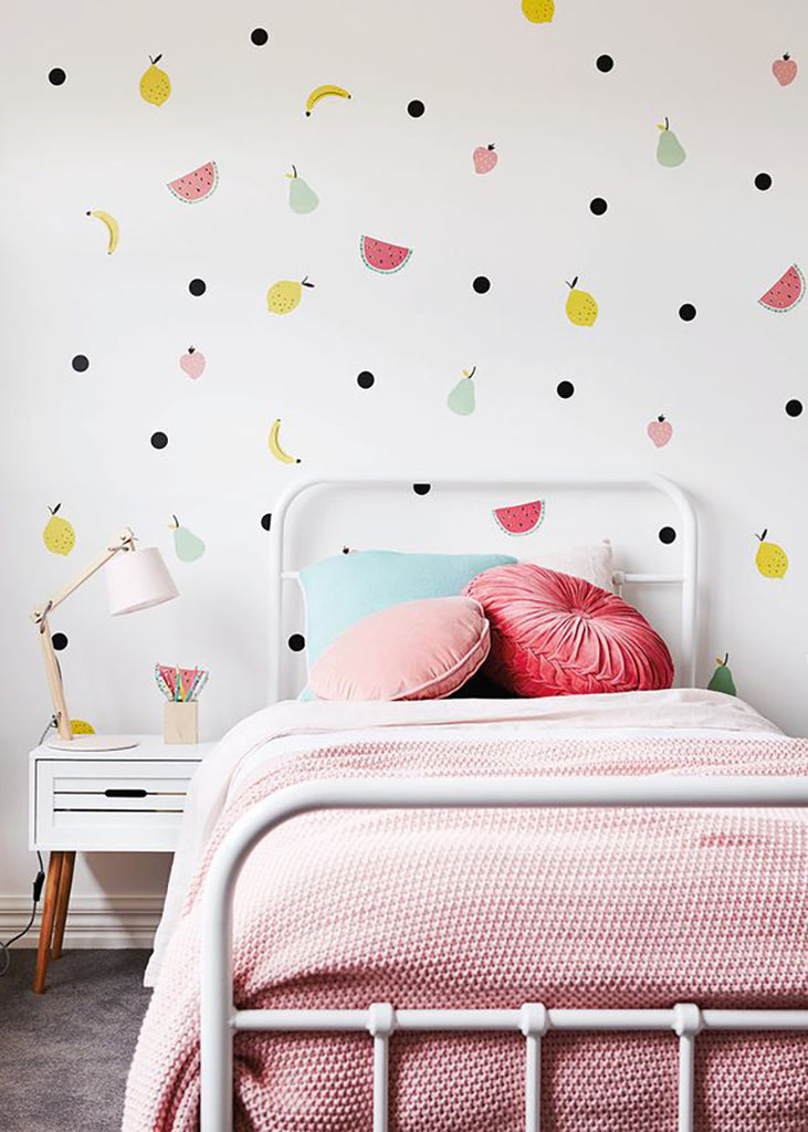 A cozy bedroom with whimsical Fruits Party, Wall Decals—stylized bananas, watermelons, and polka dots—adorning a white wall. The room also features a white bed frame with pink bedding, colorful pillows, and a nightstand with a lamp and pencils.