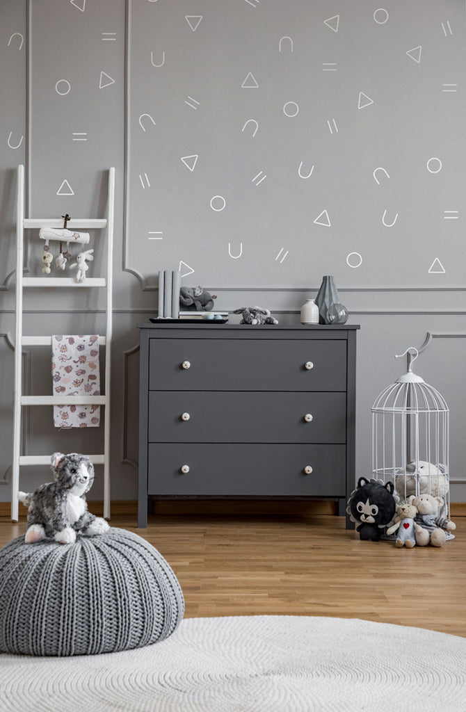 A modern room with a light grey dresser, white shelving unit, and decorative birdcage. Plush toys are arranged neatly, with a knitted pouf on a white rug. The wall features Geometric Confetti, Wall Decals in a subtle pattern.