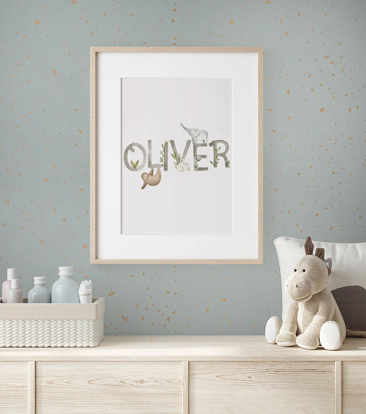 A cozy nursery features a framed print with the name "Oliver" in playful animal-themed letters on a wall adorned with Gold Metallic Confetti Speckles, Pattern Wallpaper Blue. The wooden dresser below holds neatly arranged baby essentials in a woven basket, alongside a soft, plush toy.