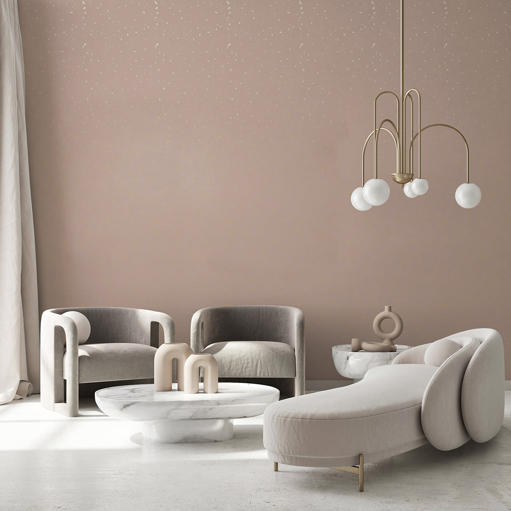 A stylish living room features modern furniture against a wall adorned with Gold Metallic Confetti Speckles, Pattern Wallpaper Pink . The space includes two sleek, curved grey armchairs, a plush, cream chaise lounge, and a round marble coffee table. A contemporary chandelier with spherical lights hangs from the ceiling, and minimalist decor items add to the elegant aesthetic. Light curtains frame the room, creating a soft, inviting atmosphere.