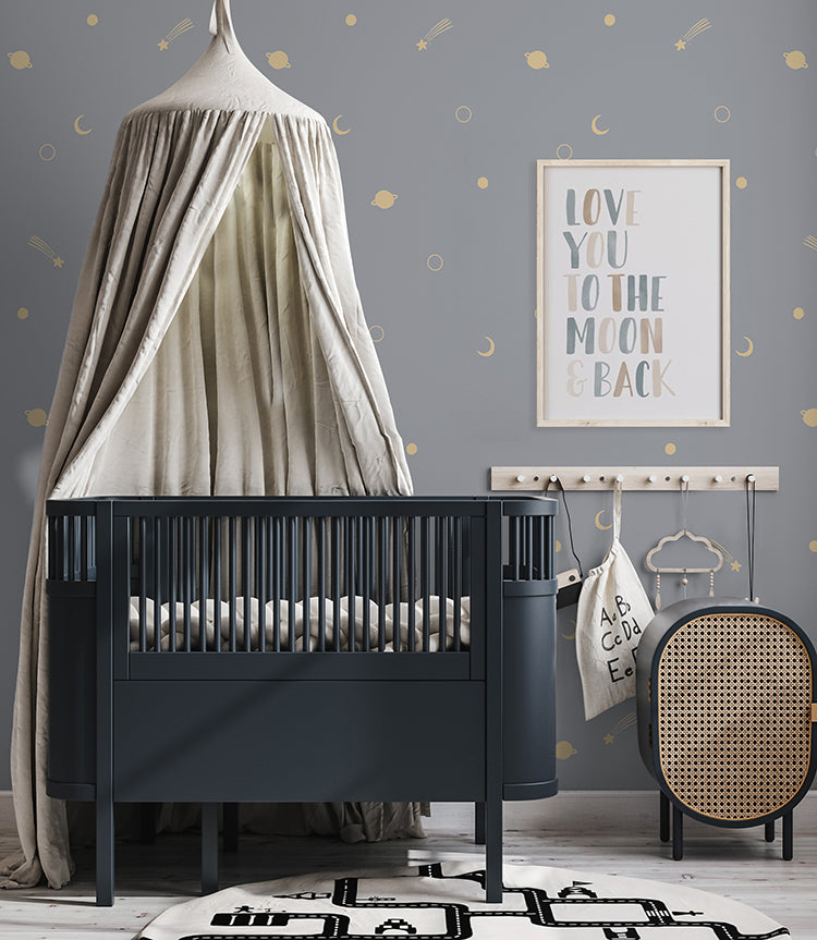 A softly lit nursery decorated with a canopy crib, a framed print saying Love you to the moon & back, and the Gold Metallic Space Galaxy, Wallpaper in Blue. The neutral blue color palette of the wallpaper and furniture creates a calming atmosphere in the room.