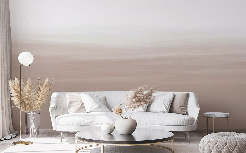 A chic living room exudes tranquility with its minimalist decor, featuring a sleek white sofa set against a subtle gradient ombre mural, titled Gradient Ombre, Mural Wallpaper in Nude. The room’s ambiance is enhanced by the soft glow of a contemporary floor lamp, while a round coffee table anchors the space, adorned with delicate pampas grass that adds a touch of organic elegance.