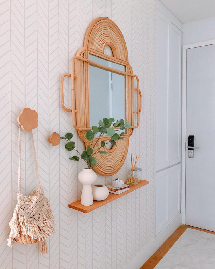 A cozy corner with soft, neutral Hand-Drawn Herringbone Pattern Wallpaper. An oval rattan mirror hangs above a wooden shelf adorned with a potted plant, white vases, and decorative items. A macrame bag hangs to the left.