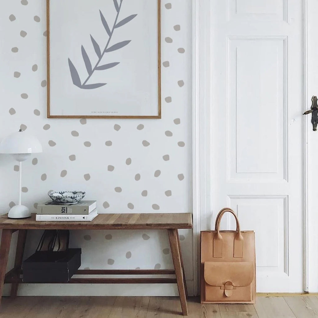 Minimalist room featuring Hand Drawn Dots, Wallpaper, a pattern of irregular beige dots on a white background. Decor includes a wooden desk, white lamp, framed leaf art, and a tan leather bag.