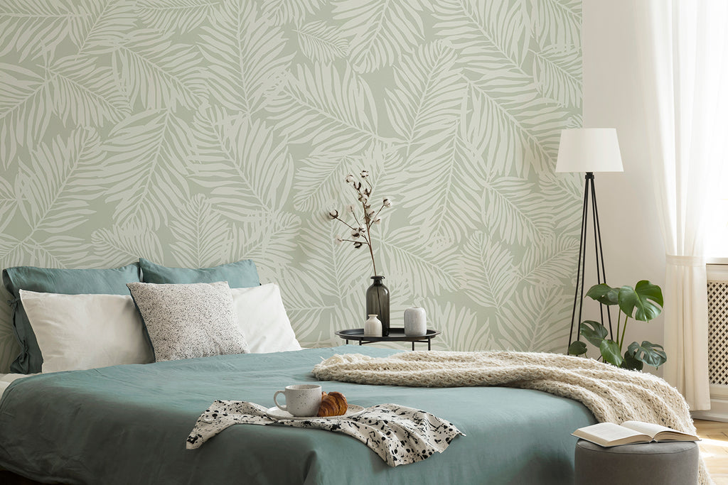 A tranquil bedroom featuring the Heather Fern, Pattern Wallpaper in Green, displaying oversized, stylized fern leaves in a calming green shade. The room houses a bed with teal and white bedding, a petite table with ornamental items, and a floor lamp.