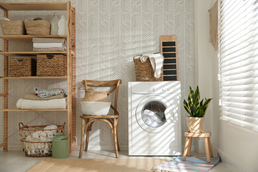 A warm, inviting laundry room adorned with Herringbone by Sophie, Pattern Wallpaper. Sunlight illuminates wooden shelves with towels, a cozy wicker chair, a modern washing machine, and lush green plants.