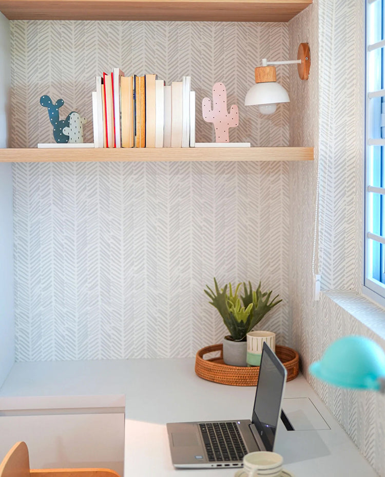 A tastefully decorated workspace with Herringbone by Sophie, Pattern Wallpaper. The room features a desk with a laptop, a shelf filled with books and cacti, and a lush plant in a wicker basket.