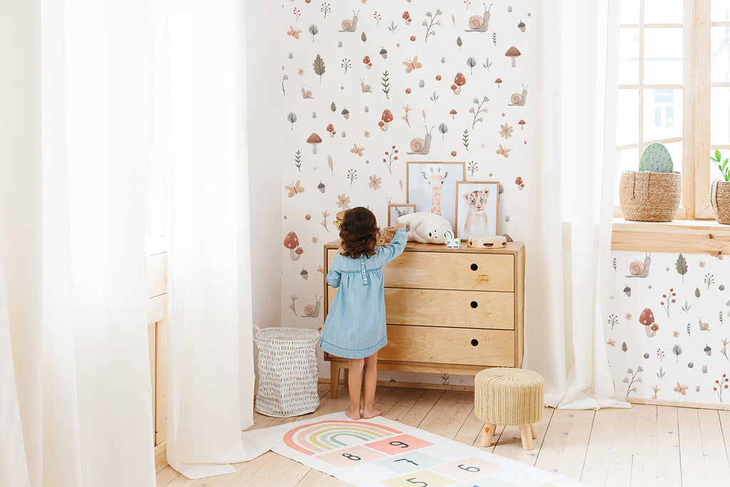 A lively child in a blue dress is in a room with ‘Lil’ Garden and Friends, Pattern Wallpaper. The room is filled with a wooden dresser, a rattan basket, and a vibrant rug, creating a playful and charming atmosphere.