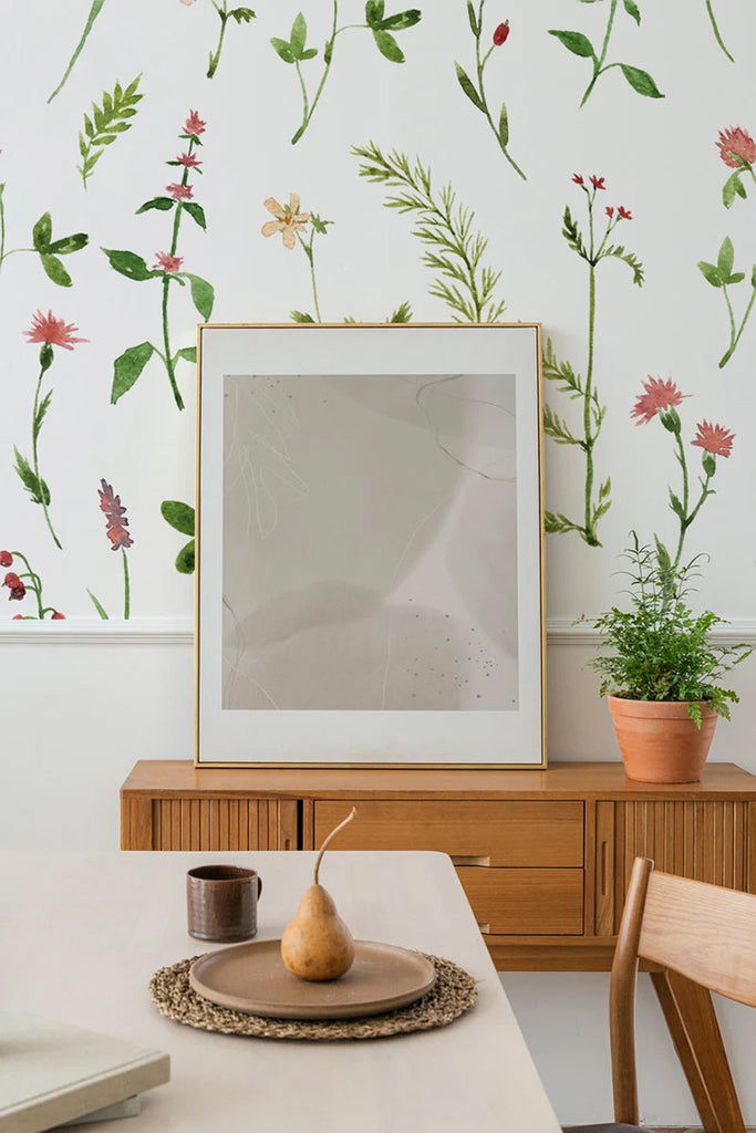 Interior with Lila, Floral Pattern Wallpaper. Wallpaper features green leaves, red flowers. A wooden sideboard holds a framed print, a plant in a terracotta pot, and a dining setup with a pear.