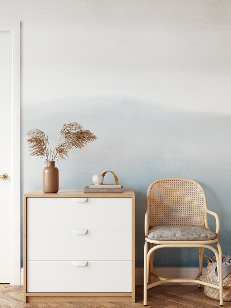 A minimalist interior features a white wooden dresser with a tan vase of dried grass, a book, and a white sphere on top. A light brown rattan chair is placed beside the dresser, and the wall behind is adorned with Marigold Mountain, Watercolor Wallpaper in Blue.