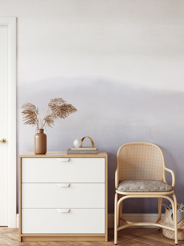 A minimalist interior features a white wooden dresser with a tan vase of dried grass, a book, and a white ornament  on top. A light brown rattan chair is placed beside the dresser, and the wall behind is adorned with Marigold Mountain, Watercolor Wallpaper in Purple.