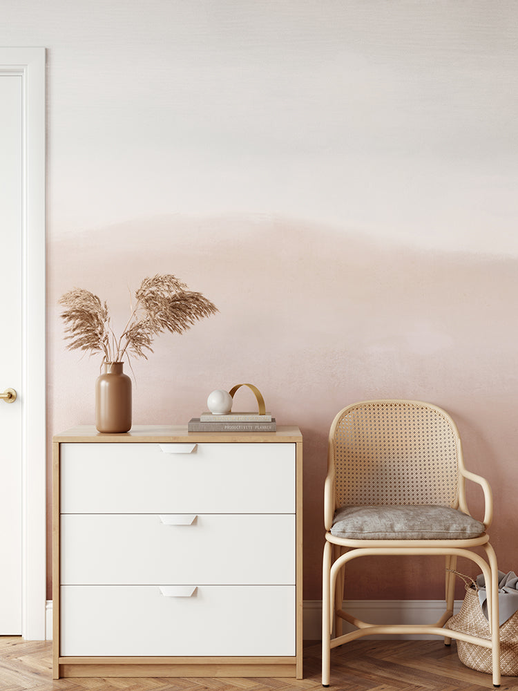 A minimalist interior features a white wooden dresser with a tan vase of dried grass, a book, and a white ornament on top. A light brown rattan chair is placed beside the dresser, and the wall behind is adorned with Marigold Mountain, Watercolor Wallpaper in Terracotta.