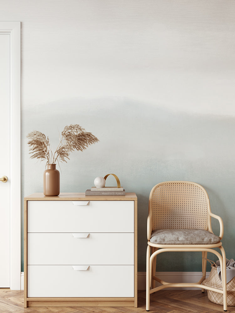 A minimalist interior features a white wooden dresser with a tan vase of dried grass, a book, and a white sphere on top. A light brown rattan chair is placed beside the dresser, and the wall behind is adorned with Marigold Mountain, Watercolor Wallpaper in Turquoise.