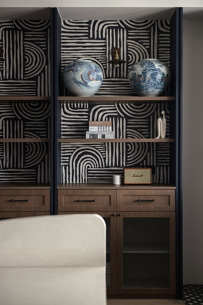 The room features dark wooden built-in shelves against a wall adorned with Midnight Brushstrokes, Pattern Wallpaper in Midnight Blue. Blue and white ornate vases and books add to the modern decor.