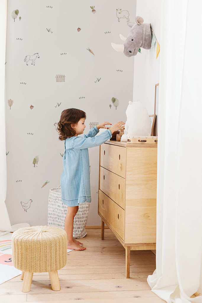 A child in a blue dress interacts with a wooden dresser against a whimsical Mini Barnyard, Pattern Wallpaper  featuring various farm animals. A plush elephant head adorns the wall, enhancing the playful theme.