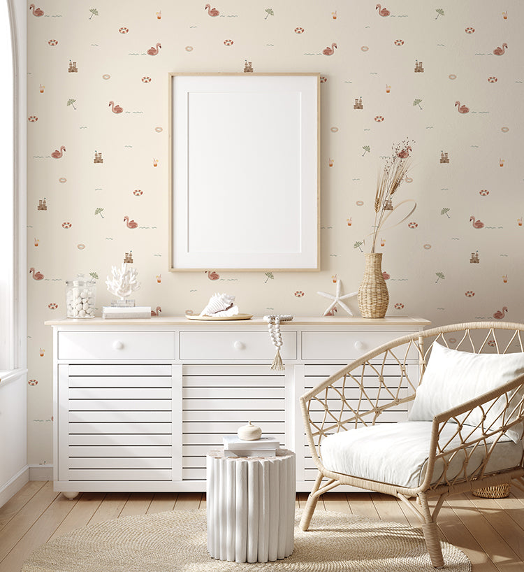 A white wooden dresser with slatted drawer fronts adds a touch of coastal charm. The dresser is accessorized with a stack of books, a vase with dried plants, and seashells. A woven armchair with a white cushion complements the natural elements of the decor along with the Mini Beach Day, Pattern Wallpaper in Sand.