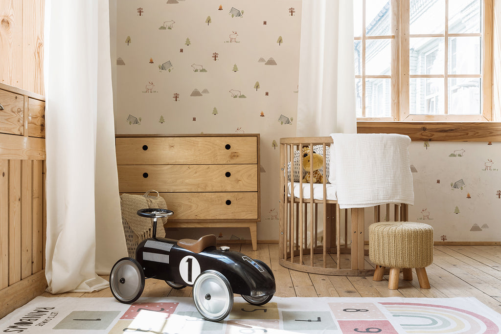 A cozy nursery features a light wood dresser and a round crib, creating a warm, inviting atmosphere. A black toy car sits on a colorful n rug, adding a playful touch. The room is accented by whimsical Mini Camper, Pattern Wallpaper in Sand, enhancing its charming, rustic feel. White curtains and ample natural light complete the serene, well-designed space