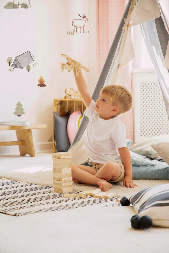 A young boy sits on a cozy, patterned rug, playing with a wooden airplane and building blocks in a beautifully designed room. A mini camper tent adds a playful touch, complemented by light-colored, wooden furniture. The soft tones and whimsical Mini Camper, Pattern Wallpaper in White decor create a charming, child-friendly space.