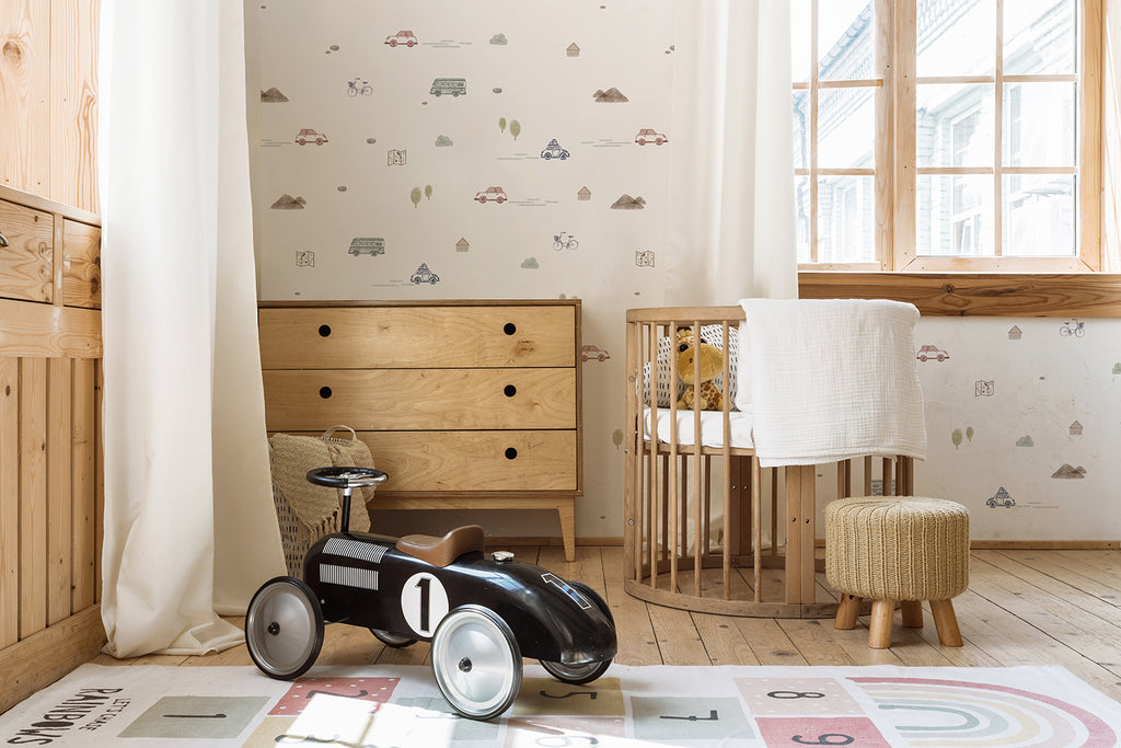 A cozy nursery features a Mini Roadtrippers, Pattern Wallpaper in Grey with whimsical vehicles traveling across the wall. The neutral color palette and wooden furniture complement the adventurous wallpaper. A black vintage toy car rests on a colorful playmat, hinting at imaginative play. The crib, adorned with a soft blanket, adds warmth and tranquility to the space. The overall design fosters a sense of wanderlust and curiosity for a young adventurer.