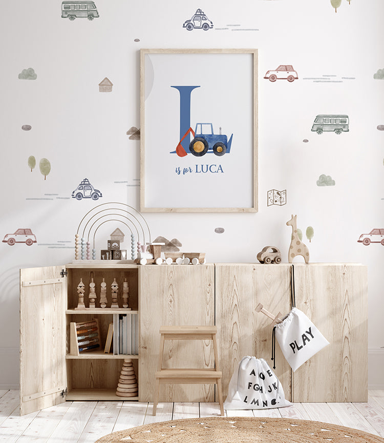 A child's playroom features a Mini Roadtrippers, Pattern Wallpaper in White showcasing colorful vehicles. A natural wood cabinet offers open storage for toys and books, with a personalized framed print above. A colorful abacus and wooden toys rest on top. A play mat on the floor provides a soft play area and complements the playful atmosphere of the room.