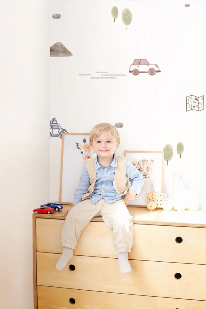 A cheerful boy sits atop a light wooden dresser in a bright, stylish room. The interior features playful decor, including framed animal prints and wooden toys. The whimsical Mini Roadtrippers, Pattern Wallpaper in White  add a touch of adventure to the clean, modern space.