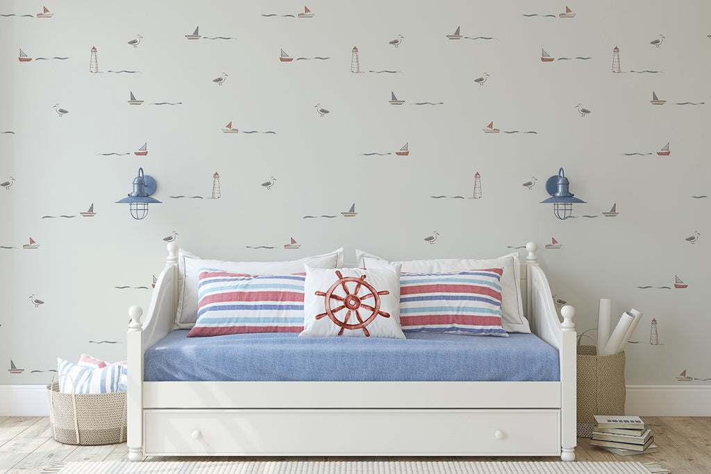 A nautical-themed room with Mini Seafarer, Pattern Wallpaper in Light Grey hues of red, blue, and beige, depicting sailboats, lighthouses, and seagulls. A white daybed with striped bedding and a maritime wheel pillow is centrally placed, accompanied by blue lantern wall sconces