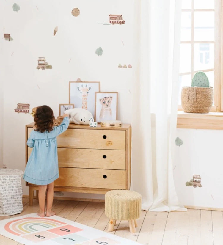 A kid’s room with a whimsical Mini Treats In The Park, Pattern Wallpaper in White. A wooden dresser with a stuffed toy, a woven basket, and a vibrant rug add to the room’s charm. A child in a blue dress adds a lively touch.