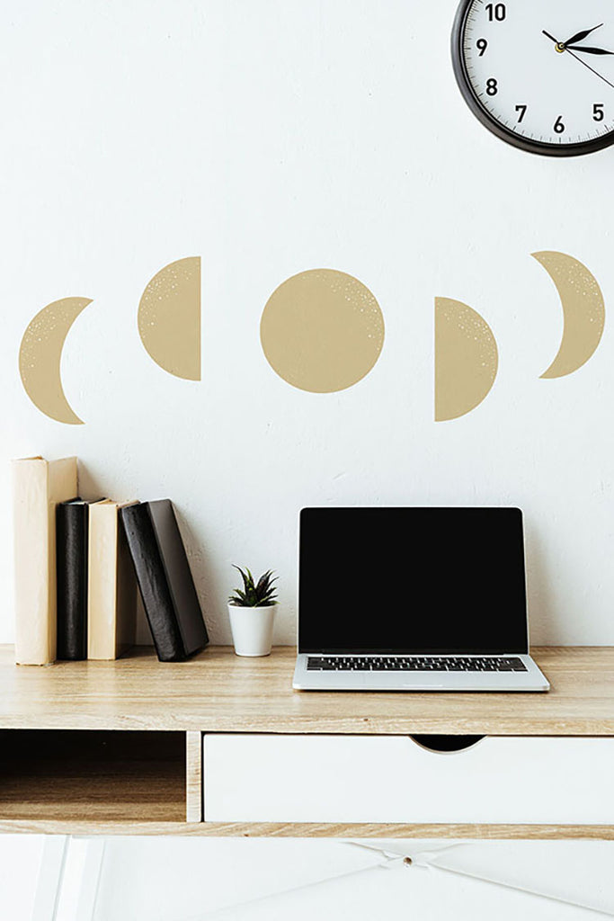 A minimalist workspace with a white desk adorned by Moon Phases, Wall Decals in Yellow. A laptop, books, and a small potted plant sit on the desk, while a wall clock hangs above.