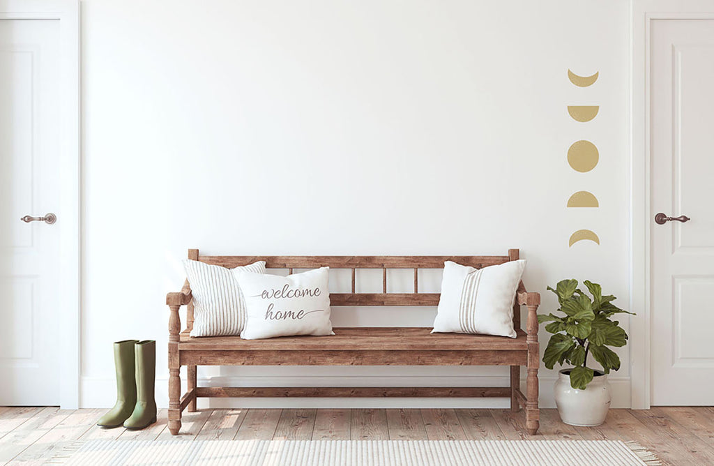 A cozy room with a wooden bench, white cushions, and a ‘welcome home’ pillow. Green rain boots rest beside the bench, and a potted plant adds freshness. Above, Moon Phases, Wall Decals in Yellow adorn the wall, creating a celestial touch.