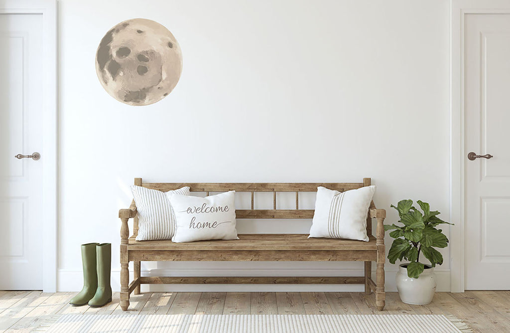 A cozy room with a wooden bench, white cushions, and a ‘welcome home’ pillow. Green rain boots rest beside the bench, and a potted plant adds freshness. Above, Moon, Wall Decals in Yellow adorn the wall, creating a celestial touch.