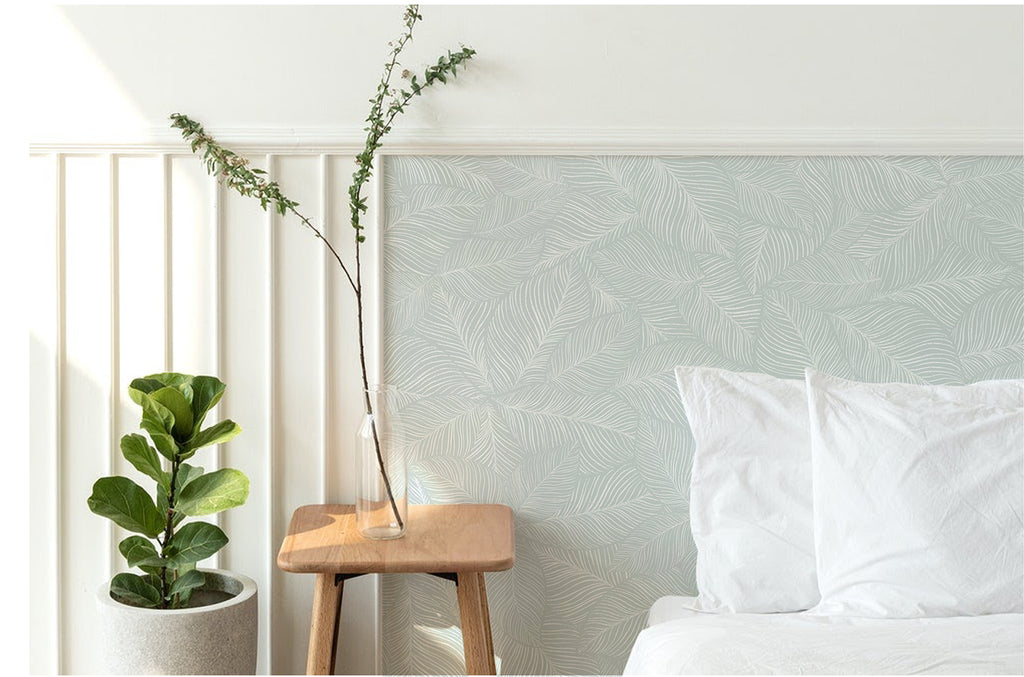 A tranquil bedroom adorned with Noelle Fern, Tropical Pattern Wallpaper in Blue. The subtle leaf design enhances the room’s serene ambiance. A cozy white bed and a wooden stool with a potted plant complete the scene.