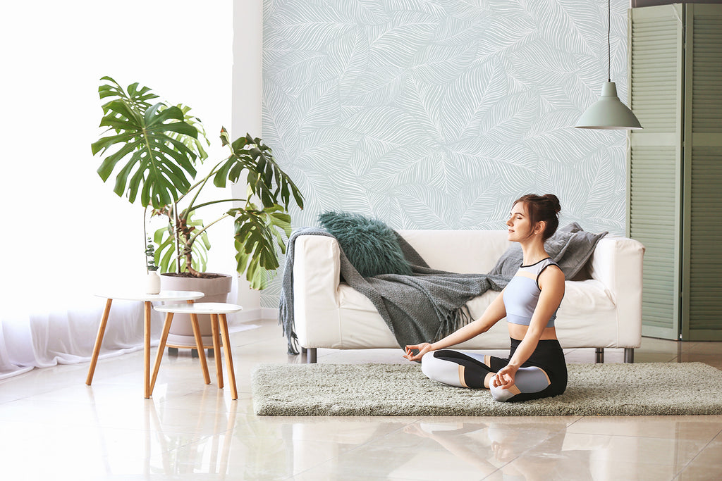 A serene room with Noelle Fern, Tropical Pattern Wallpaper in Blue. A person in a yoga pose on a mat, facing a white sofa with a blue throw. A wooden table and potted plant add natural elements.