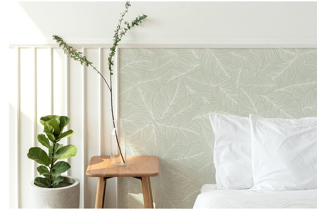 A tranquil bedroom adorned with Noelle Fern, Tropical Pattern Wallpaper in Green. The subtle leaf design enhances the room’s serene ambiance. A cozy white bed and a wooden stool with a potted plant complete the scene.