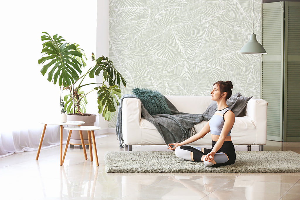 A tranquil bedroom adorned with Noelle Fern, Tropical Pattern Wallpaper in Green. The subtle leaf design enhances the room’s serene ambiance. A cozy white bed and a wooden stool with a potted plant complete the scene.