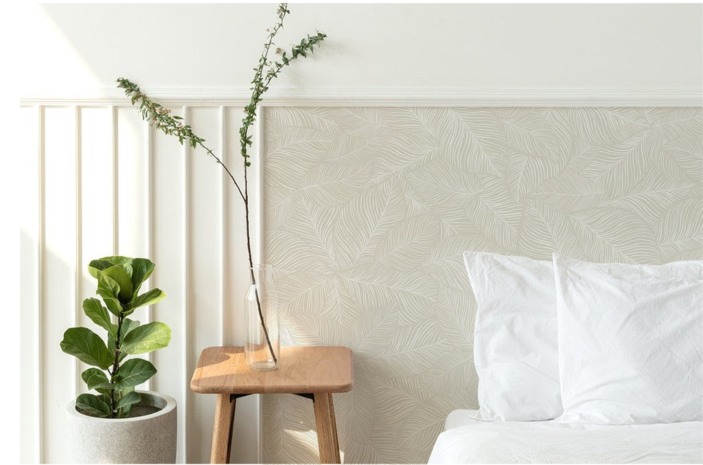 A tranquil bedroom adorned with Noelle Fern, Tropical Pattern Wallpaper in Sand. The subtle leaf design enhances the room’s serene ambiance. A cozy white bed and a wooden stool with a potted plant complete the scene.