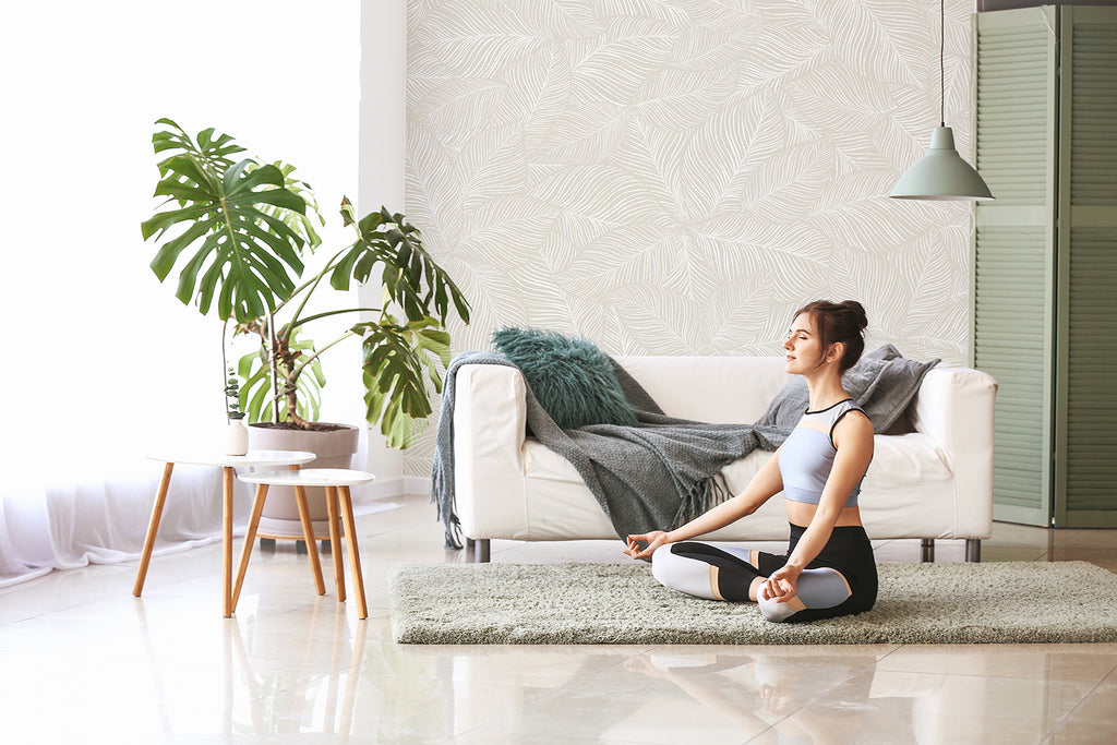 A tranquil bedroom adorned with Noelle Fern, Tropical Pattern Wallpaper in Sand. The subtle leaf design enhances the room’s serene ambiance. A cozy white bed and a wooden stool with a potted plant complete the scene.