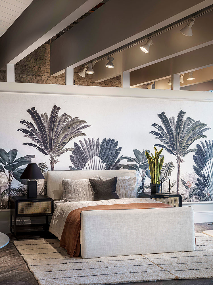 A chic bedroom adorned with Palm Paradise, Tropical Mural Wallpaper in Green, featuring large palm leaf patterns. The room includes a cozy bed with textured linens, a sleek side table, and a lush potted plant.