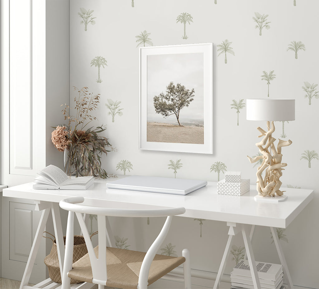 A tranquil workspace adorned with Pamela Palm, Tropical Pattern Wallpaper in Green. A white desk with an open book, a unique lamp, and decor items, under a framed tree picture.