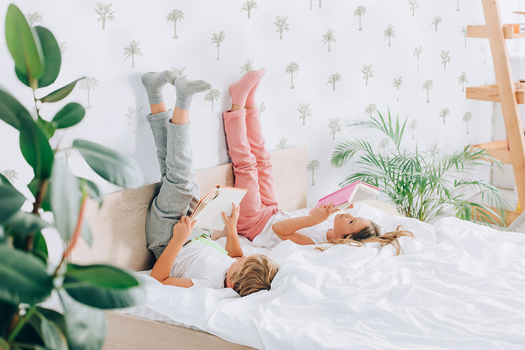 Two individuals relax on a bed, reading books with legs up against a wall. The wall is adorned with Pamela Palm, Tropical Pattern Wallpaper in Green, adding a tropical vibe to the room.