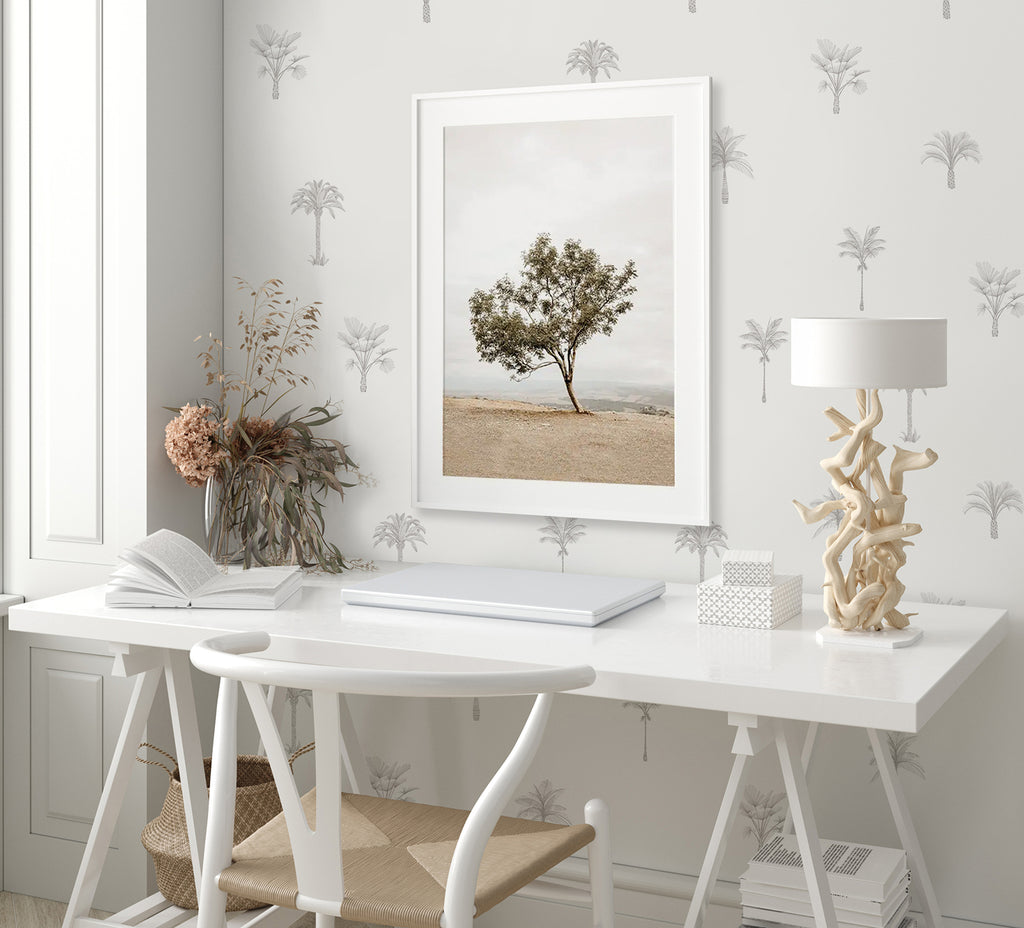 A tranquil workspace adorned with Pamela Palm, Tropical Pattern Wallpaper in Grey. A white desk with an open book, a unique lamp, and decor items, under a framed tree picture.