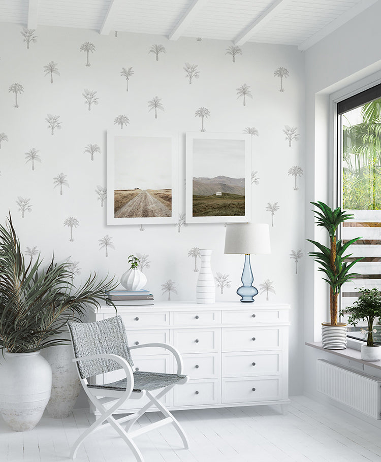 A vibrant room with Pamela Palm, Tropical Pattern Wallpaper in Grey. White furniture, a rocking chair, and indoor plants complement the palm motifs. Framed landscapes hang above a dresser by a window.