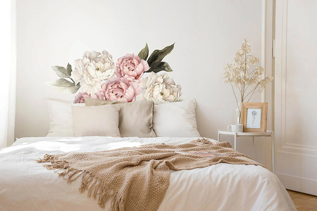 A bedroom with a calming ambiance, featuring a large wall decal of soft pink and white peonies, named Peonies Pack, Floral Wall Decals in Blush and White, above a neatly made bed with white bedding and a tan throw.