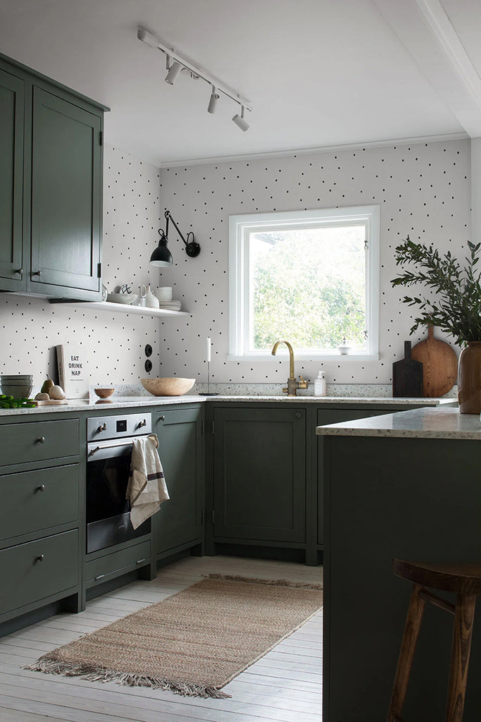 A cozy kitchen with olive green cabinets and Polka Delicate Dots, Wallpaper in Black White. A window above the sink lets in natural light, highlighting the wooden countertops and beige rug.
