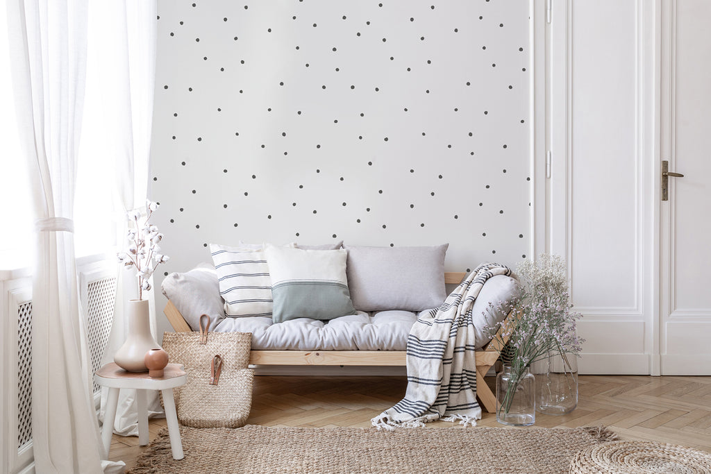 An elegant interior with a white sofa, cushions, and a cozy blanket. The wall features a Polka Delicate Dots, Wallpaper in Black White. A wicker basket, decorative vase with branches, and wooden floor add charm.