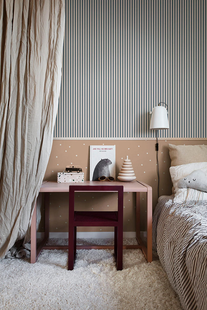 A cozy bedroom corner with Polka Delicate Dots, Wallpaper in Nude. A striped wall complements the dot pattern. A draped beige curtain, wooden bedside table, and zebra-striped bedding add texture.