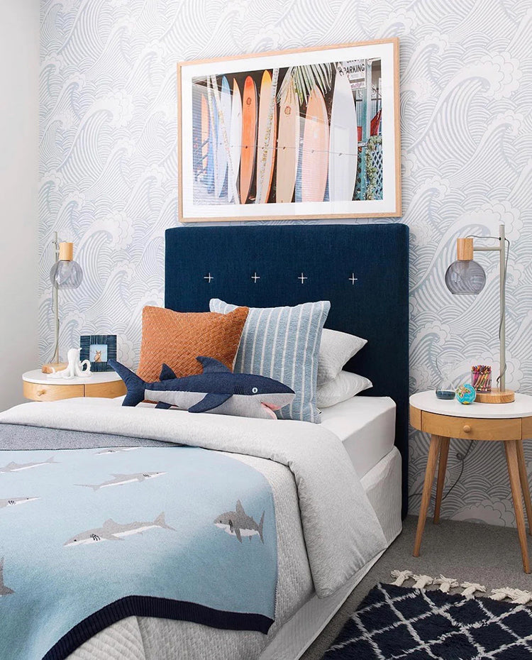 A serene bedroom featuring Sail Away, Blue Pattern Wallpaper with stylized waves and sailboats. A navy blue headboard contrasts with white maritime-themed bedding. Nautical artwork completes the theme.